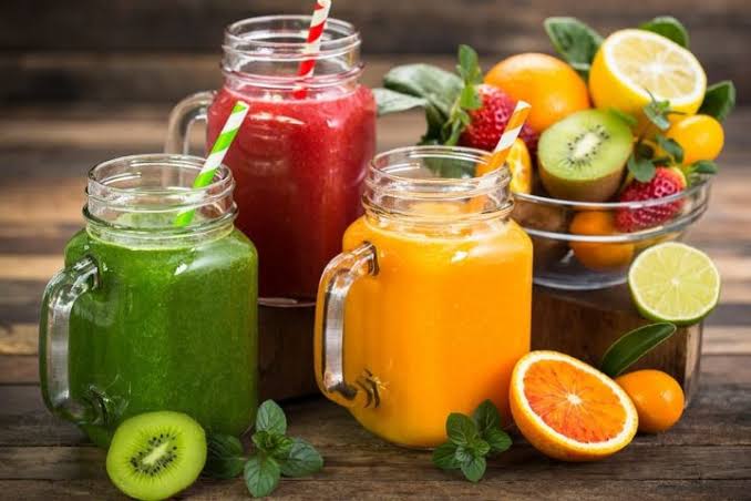 Copos de sucos naturais coloridos de acerola, caju e maracujá dispostos sobre mesa rústica de madeira com frutas tropicais ao redor